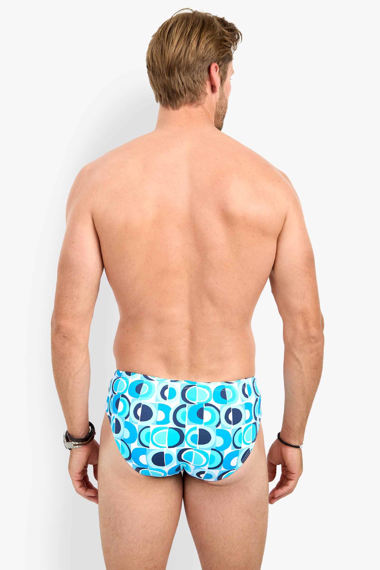 A man with light skin and short brown hair is standing with his back to the camera, wearing the BONDI SWIM BRIEF in a blue and white pattern. He has bracelets on both wrists.