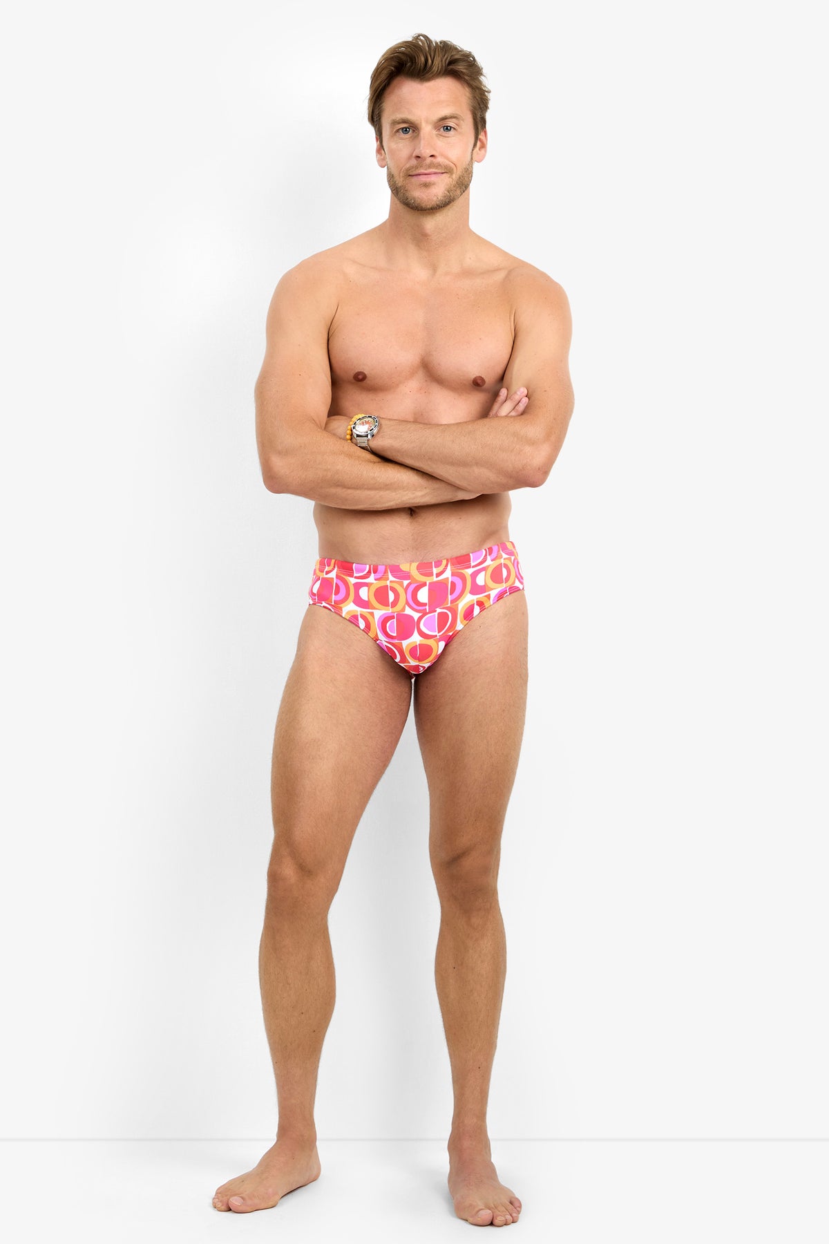 A man stands barefoot against a white background, wearing the BONDI SWIM BRIEF. His arms are crossed, and he faces the camera with a neutral expression.