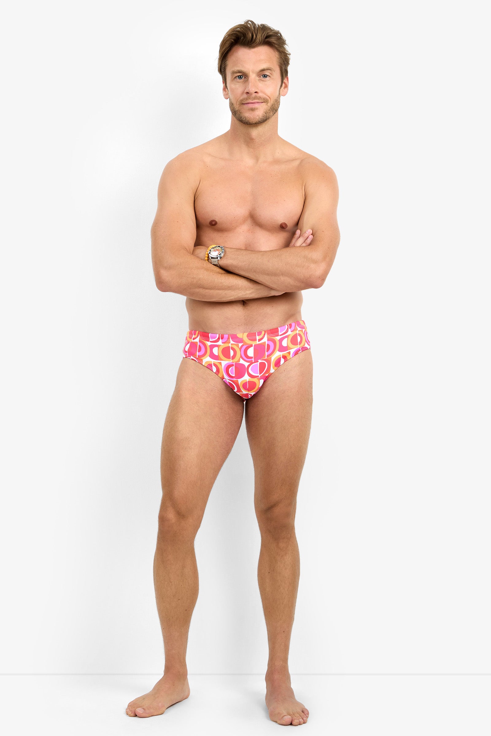 A man stands barefoot against a white background, wearing the BONDI SWIM BRIEF. His arms are crossed, and he faces the camera with a neutral expression.