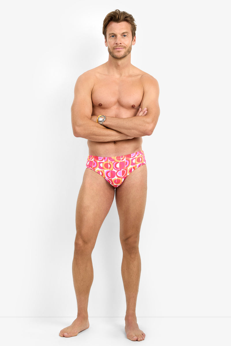 A man stands barefoot against a white background, wearing the BONDI SWIM BRIEF. His arms are crossed, and he faces the camera with a neutral expression.