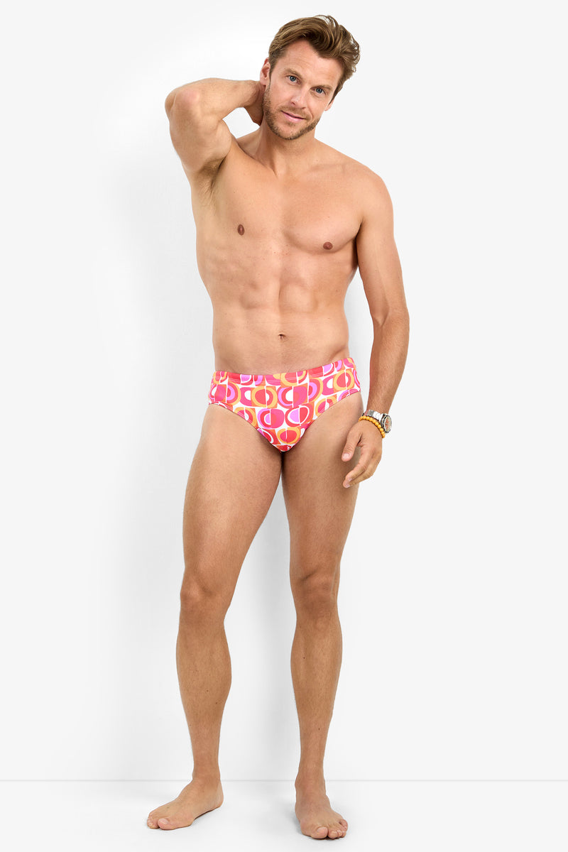 A man stands barefoot against a white background, wearing the BONDI SWIM BRIEF in a pink patterned design. He rests one hand behind his head and looks at the camera with a slight smile.