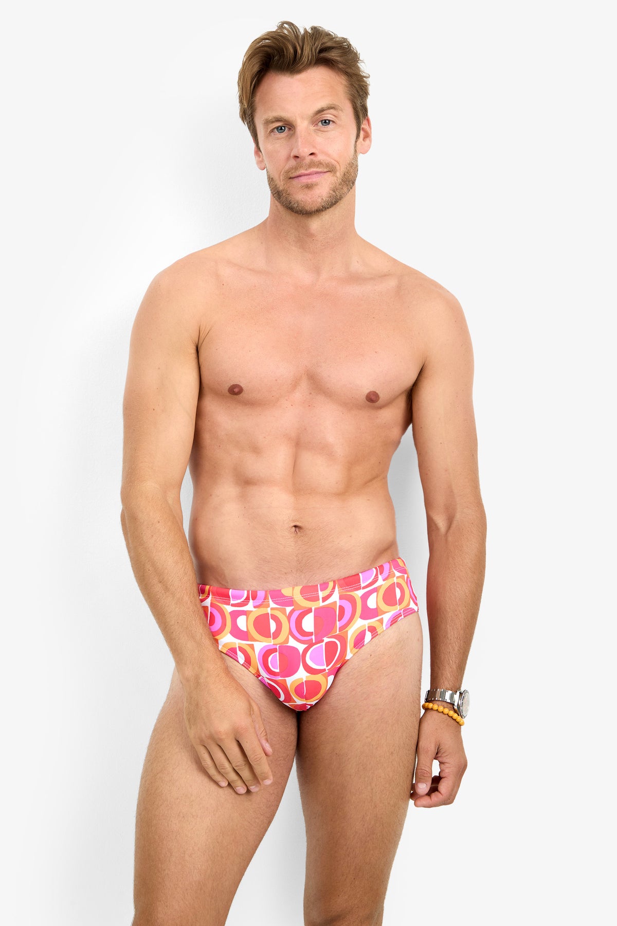 A man with light brown hair is standing against a white background, wearing the BONDI SWIM BRIEF in a pink and orange geometric pattern. He is looking at the camera and smiling slightly.