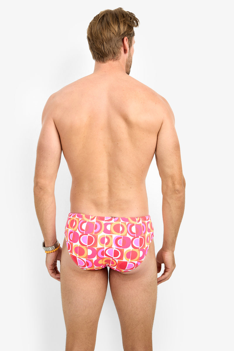 A man with light skin and brown hair stands facing away, wearing the BONDI SWIM BRIEF—pink patterned swim briefs featuring circular designs on a white background.