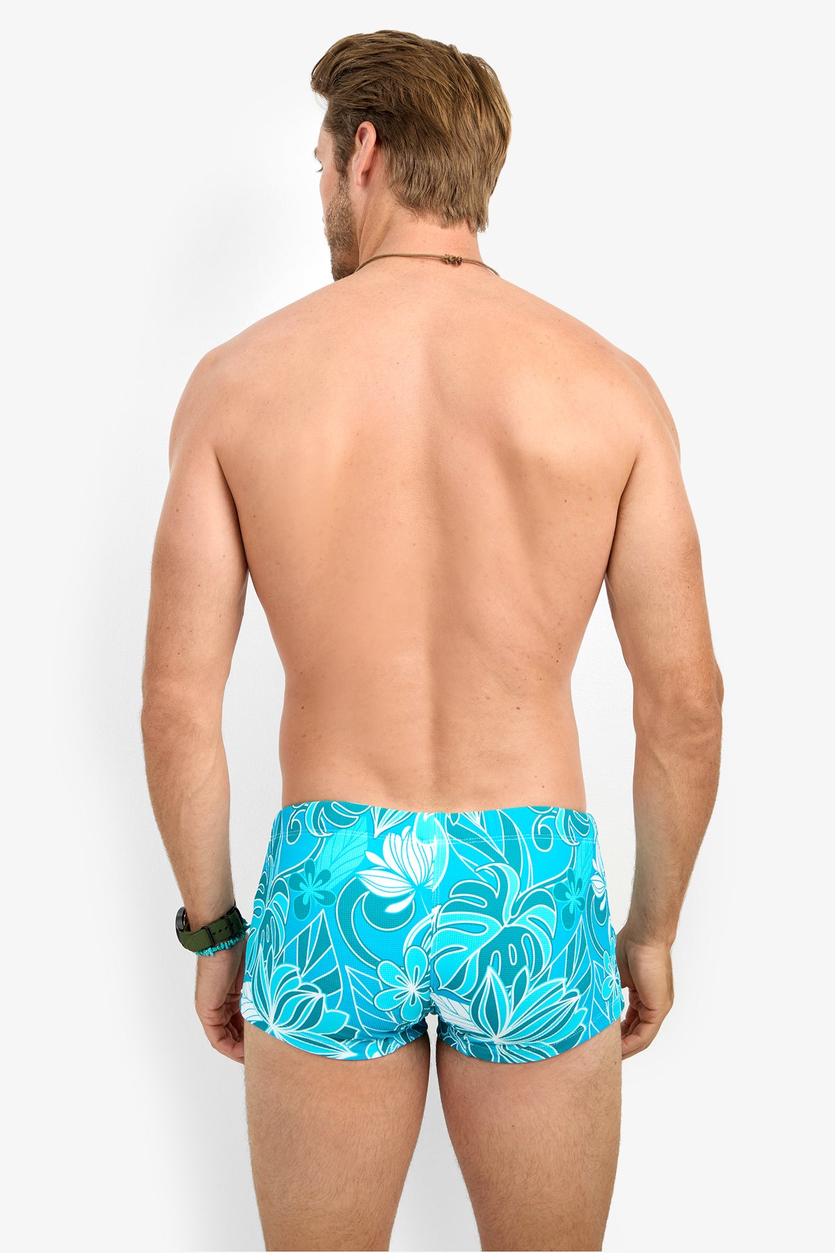 A man stands with his back to the camera, wearing MONTEGO BAY SWIM TRUNK in a bright blue floral print. His arms are relaxed by his sides, and he is shirtless against a plain white background.