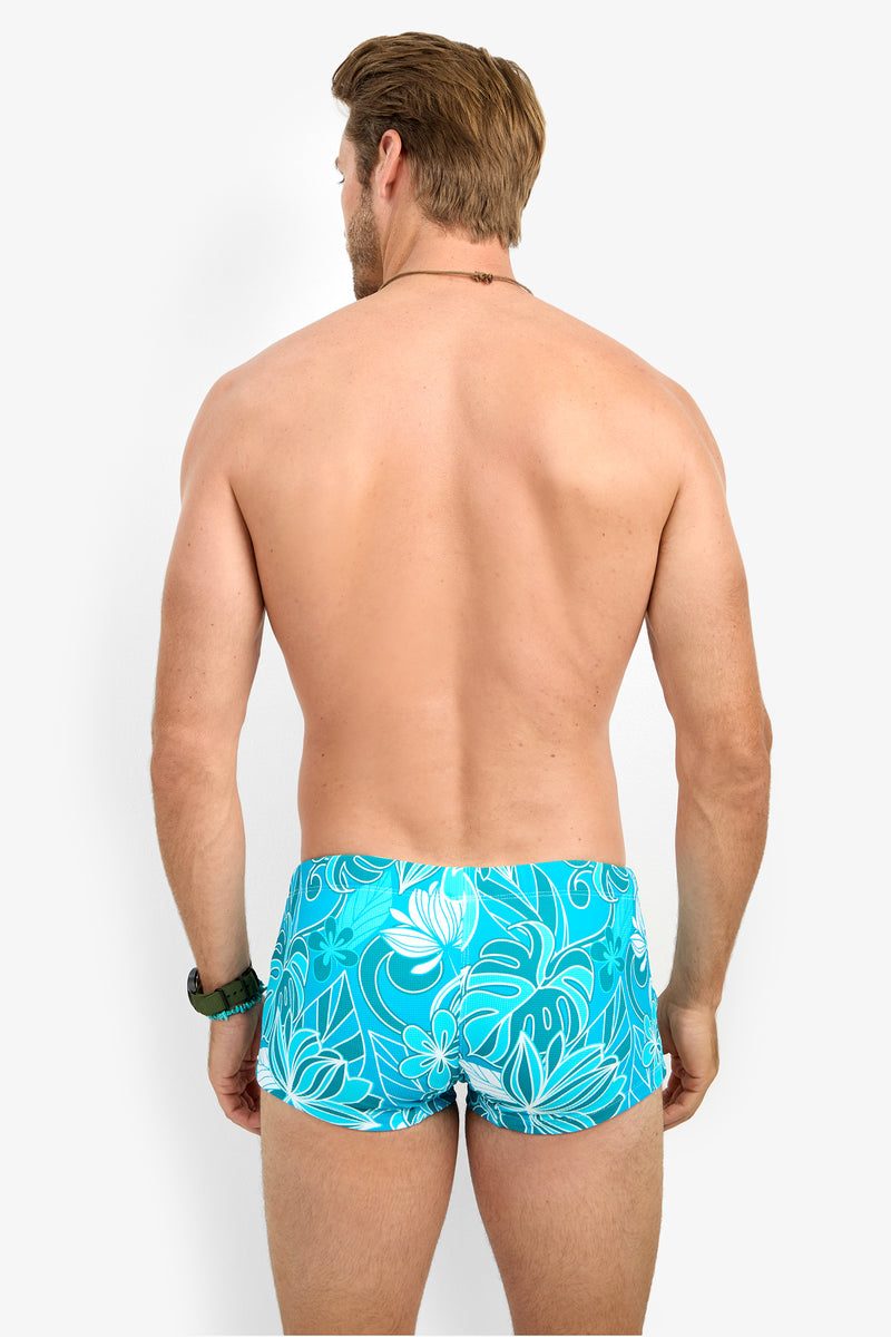A man stands with his back to the camera, wearing MONTEGO BAY SWIM TRUNK in a bright blue floral print. His arms are relaxed by his sides, and he is shirtless against a plain white background.