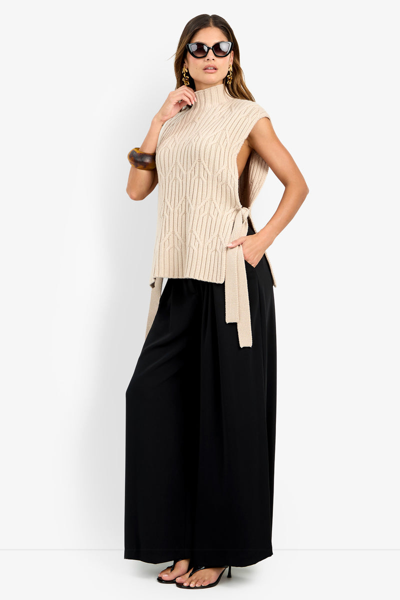A woman wearing sunglasses and the ROMEA CAPE—a sleeveless beige knit top with side ties crafted from soft wool fabric—paired with wide-leg black pants, high heels, hoop earrings, and a large bracelet, poses with one hand in her pocket against a white background.