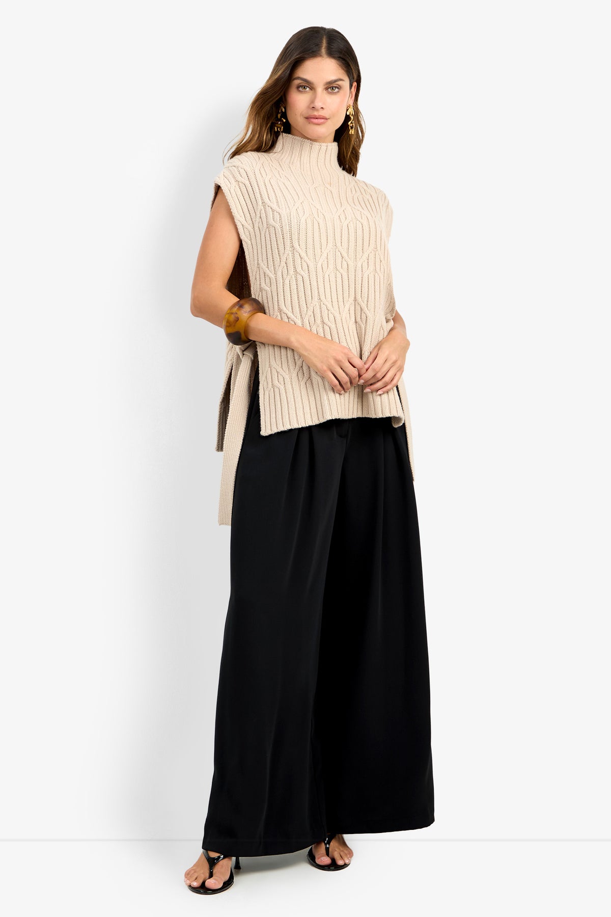 A woman wearing the ROMEA CAPE, a sleeveless beige knit sweater crafted from 100% wool with a high neck, paired with wide-leg black pants and black sandals, poses against a plain white background.