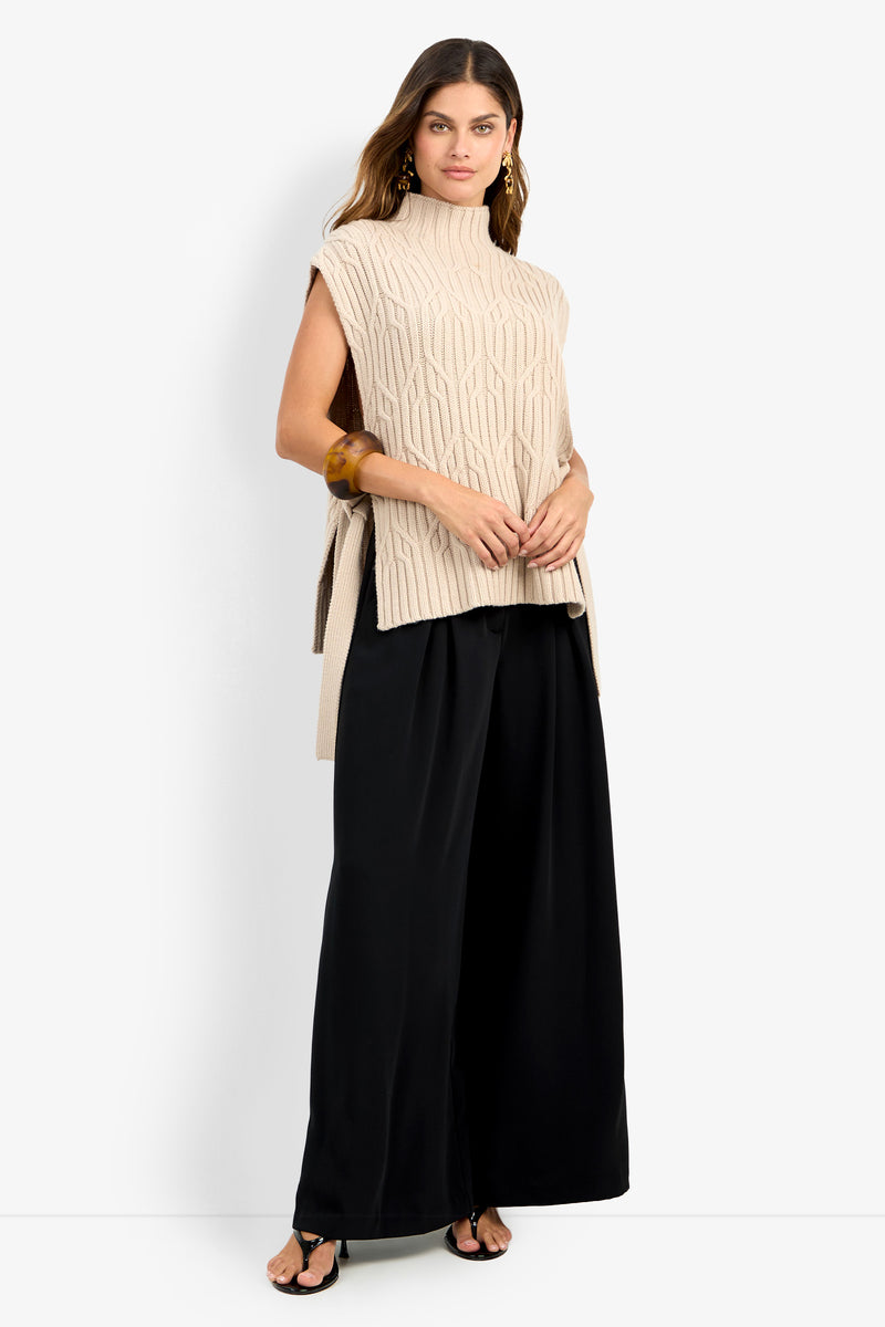 A woman wearing the ROMEA CAPE, a sleeveless beige knit sweater crafted from 100% wool with a high neck, paired with wide-leg black pants and black sandals, poses against a plain white background.