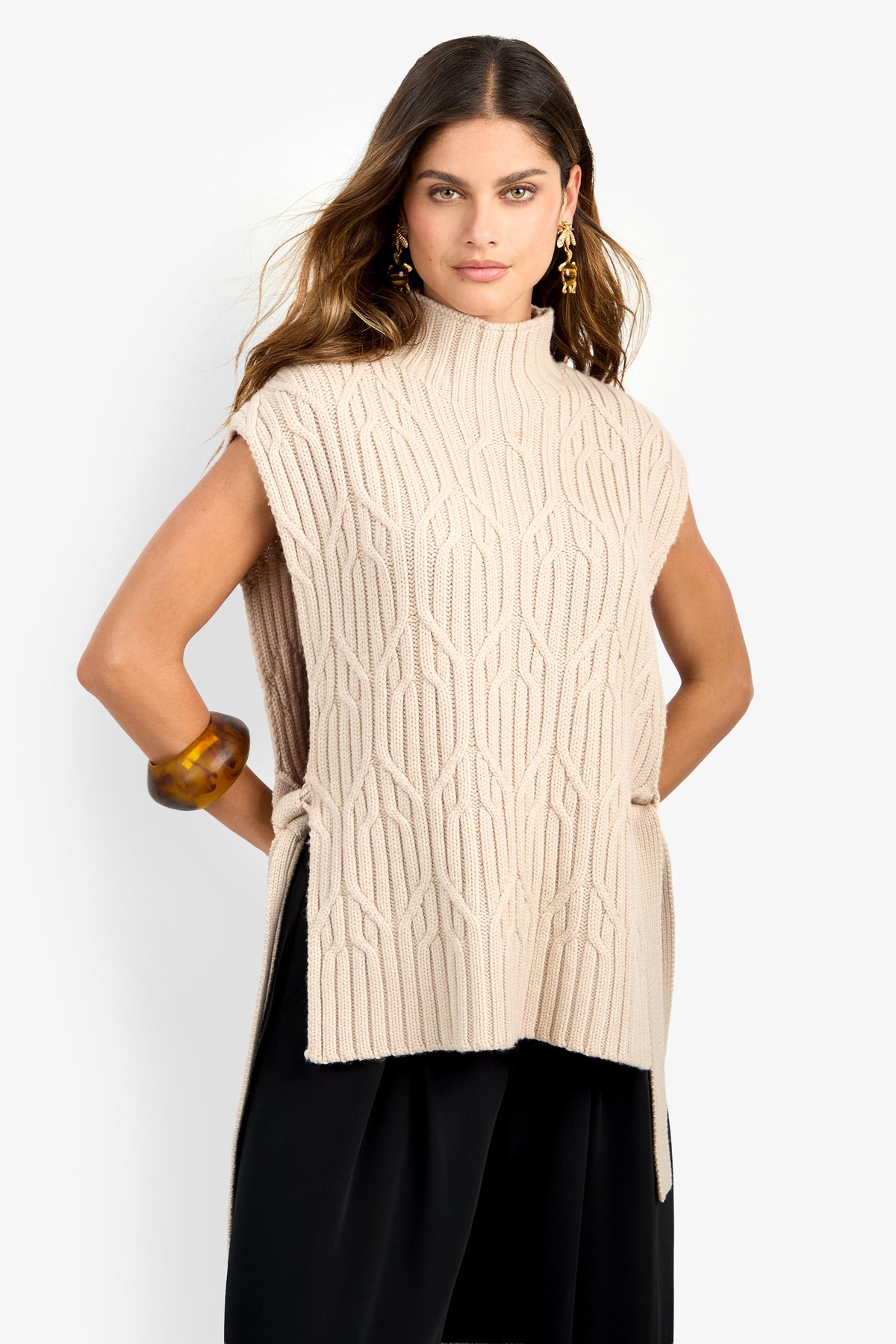 A woman stands against a plain white background, wearing the ROMEA CAPE—a sleeveless, beige textured knit sweater crafted from 100% wool with a high neck and side slits—paired with black pants, gold earrings, and a chunky brown bracelet.