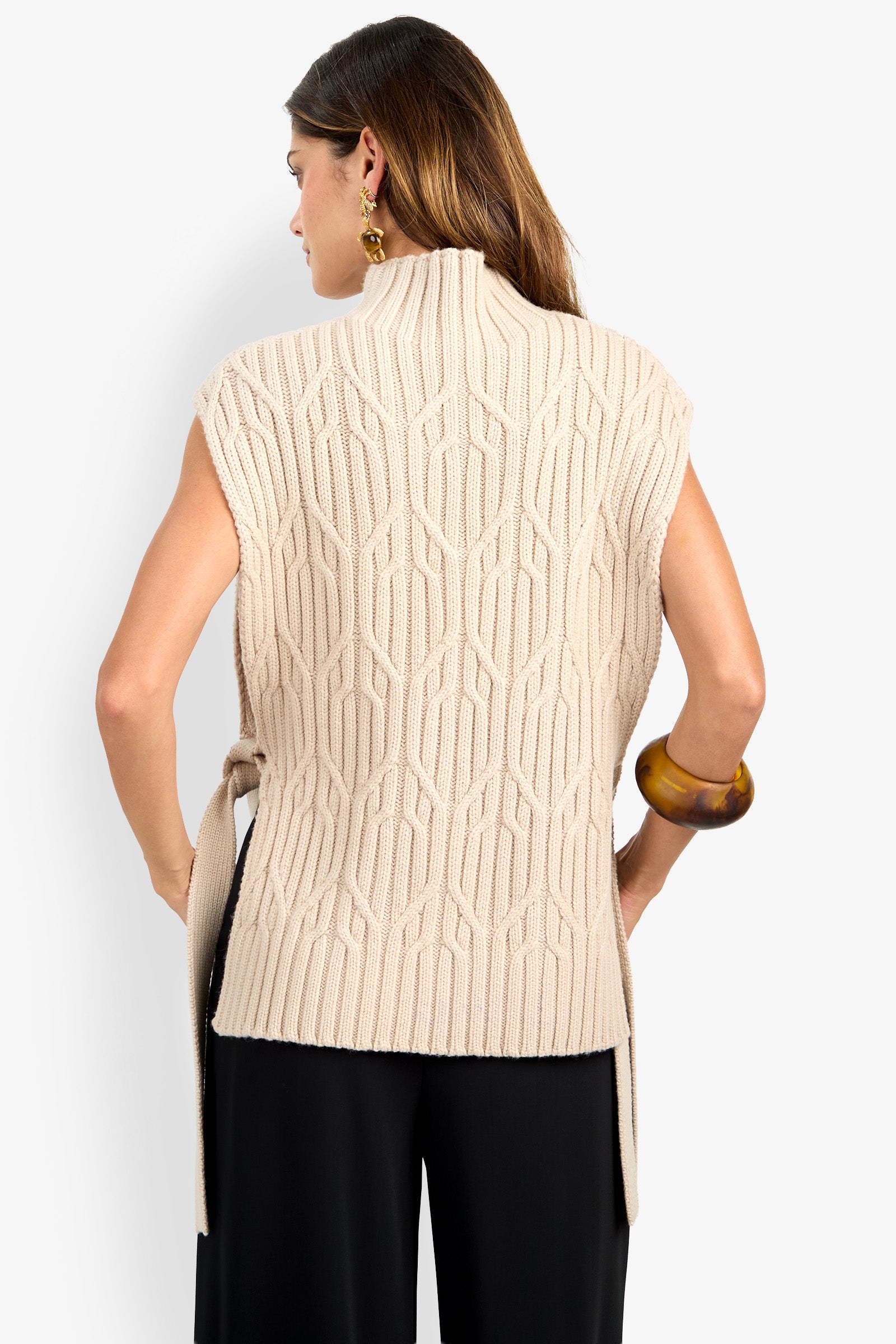 A woman is seen from behind wearing the ROMEA CAPE—a sleeveless, beige knit sweater made from soft 100% wool and featuring a geometric pattern—paired with black pants. She has brown hair and accessorizes with gold earrings and a large brown bracelet.
