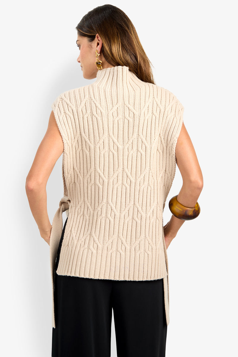 A woman is seen from behind wearing the ROMEA CAPE—a sleeveless, beige knit sweater made from soft 100% wool and featuring a geometric pattern—paired with black pants. She has brown hair and accessorizes with gold earrings and a large brown bracelet.