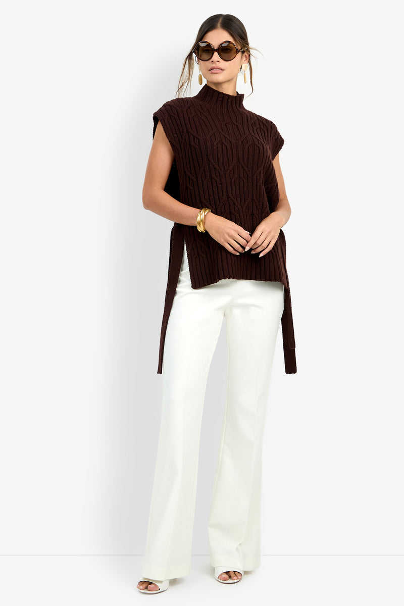Woman wearing sunglasses and the ROMEA CAPE—a sleeveless dark brown knit sweater vest crafted from 100% wool—paired with white wide-leg pants, white heeled sandals, gold hoop earrings, and a chunky gold bracelet, standing against a plain light background.