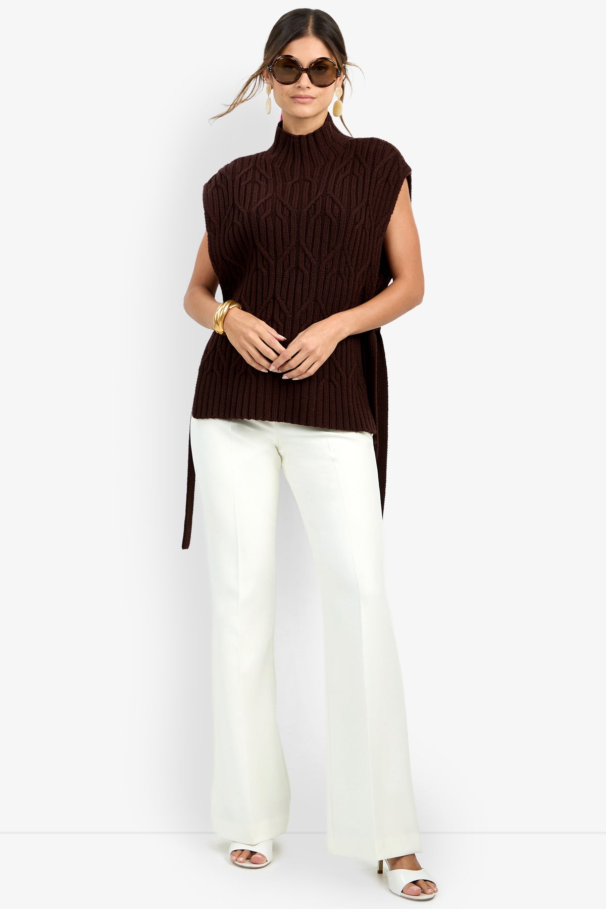 Woman wearing sunglasses, a sleeveless dark brown ROMEA CAPE knit turtleneck sweater crafted from pure wool with side slits, white wide-leg pants, white sandals, and gold jewelry, posing against a plain light background.
