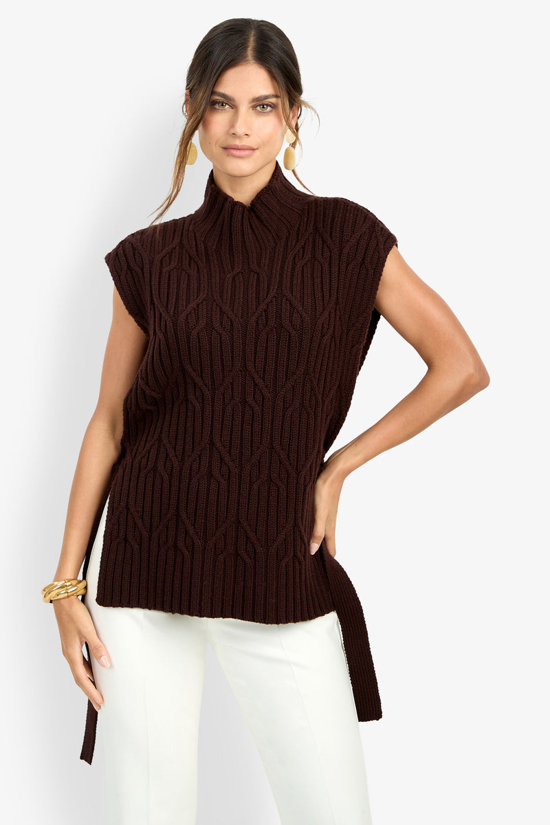 A woman models the ROMEA CAPE, a sleeveless dark brown pure wool knit top featuring a high collar and geometric pattern. She pairs it with white pants, accessorizing with gold earrings and a chunky gold bracelet, standing with one hand on her hip against a plain background.