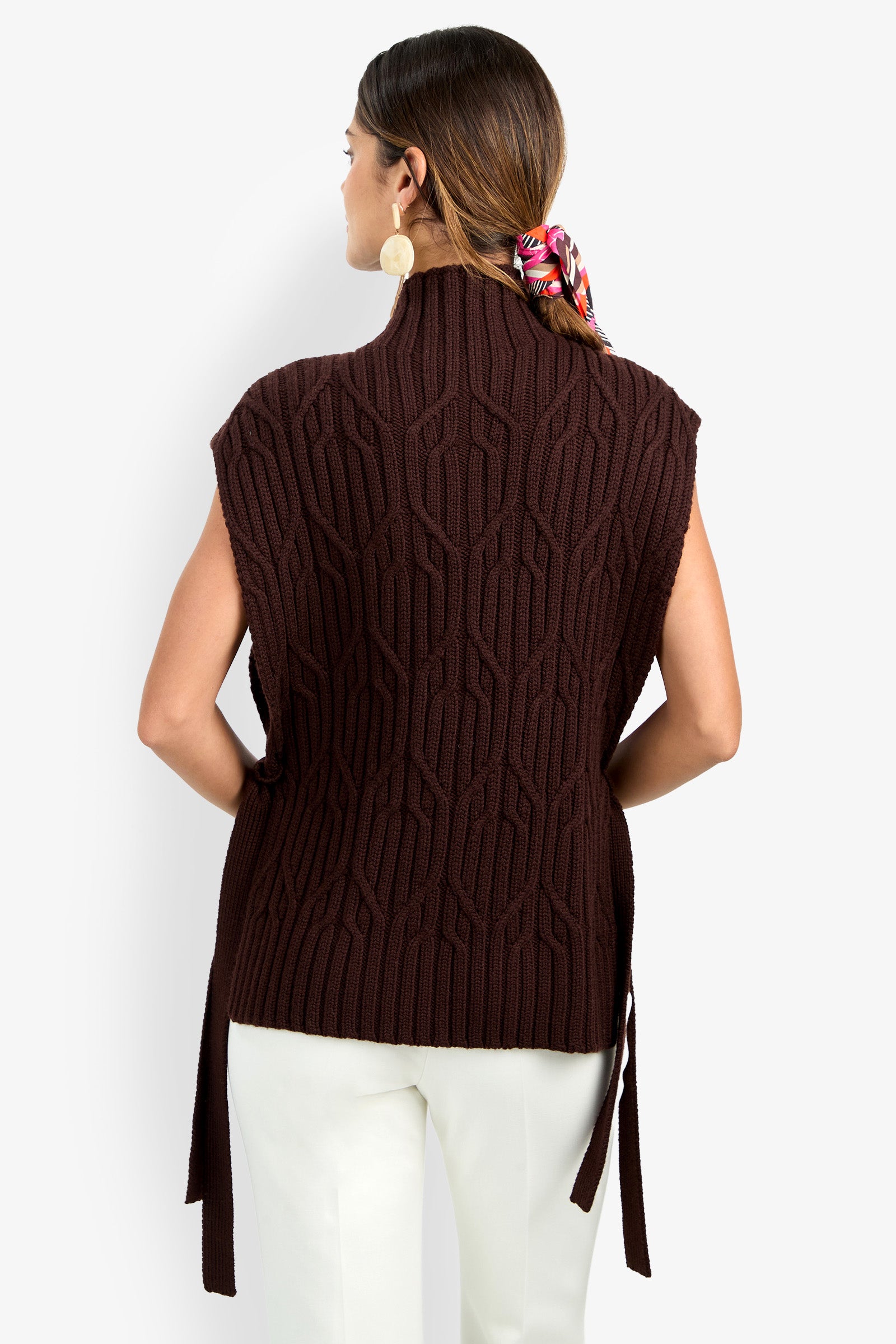 A woman with brown hair in a colorful scarf is seen from behind, wearing the ROMEA CAPE—a sleeveless, dark brown, textured pure wool sweater—paired with white pants against a plain background.