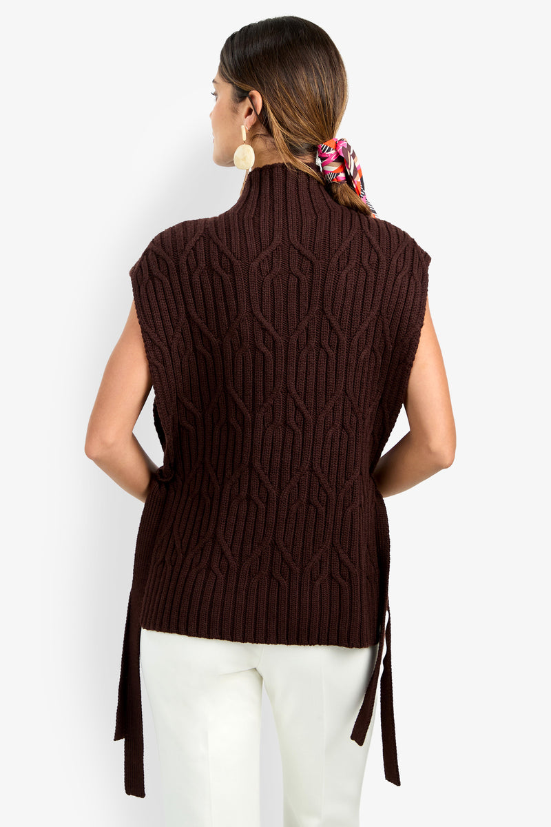 A woman with brown hair in a colorful scarf is seen from behind, wearing the ROMEA CAPE—a sleeveless, dark brown, textured pure wool sweater—paired with white pants against a plain background.