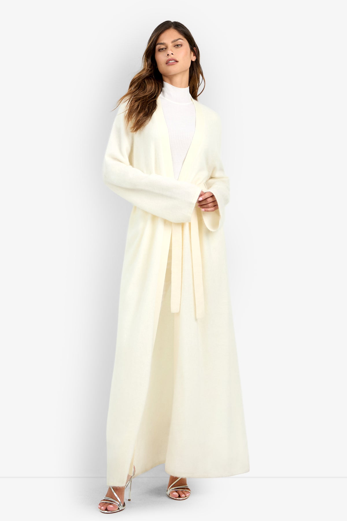A woman wears a long, cream-colored Carducci Cardigan open over a white high-neck top.