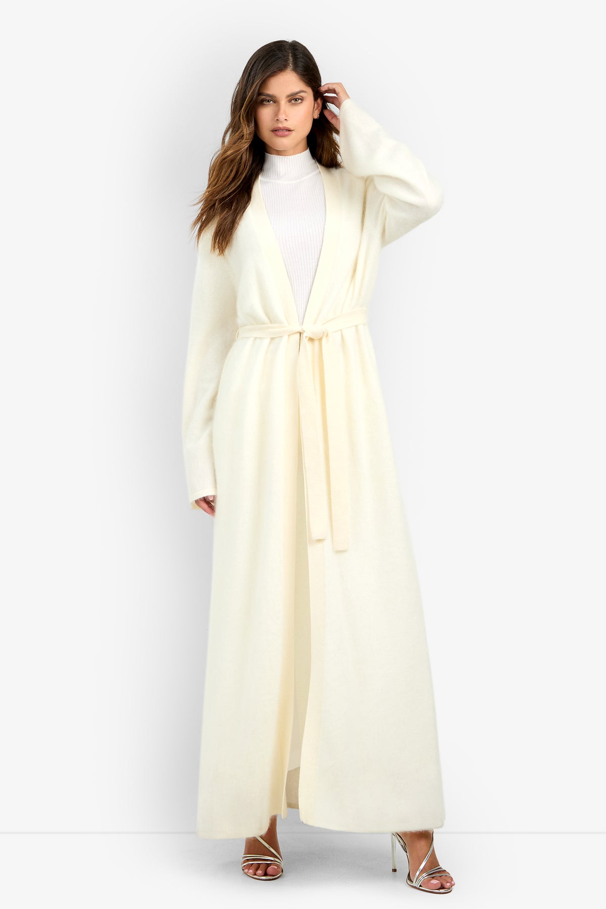 A woman wears a cream-colored, belted Carducci Cardigan over a white mock-neck top and silver strappy heels.