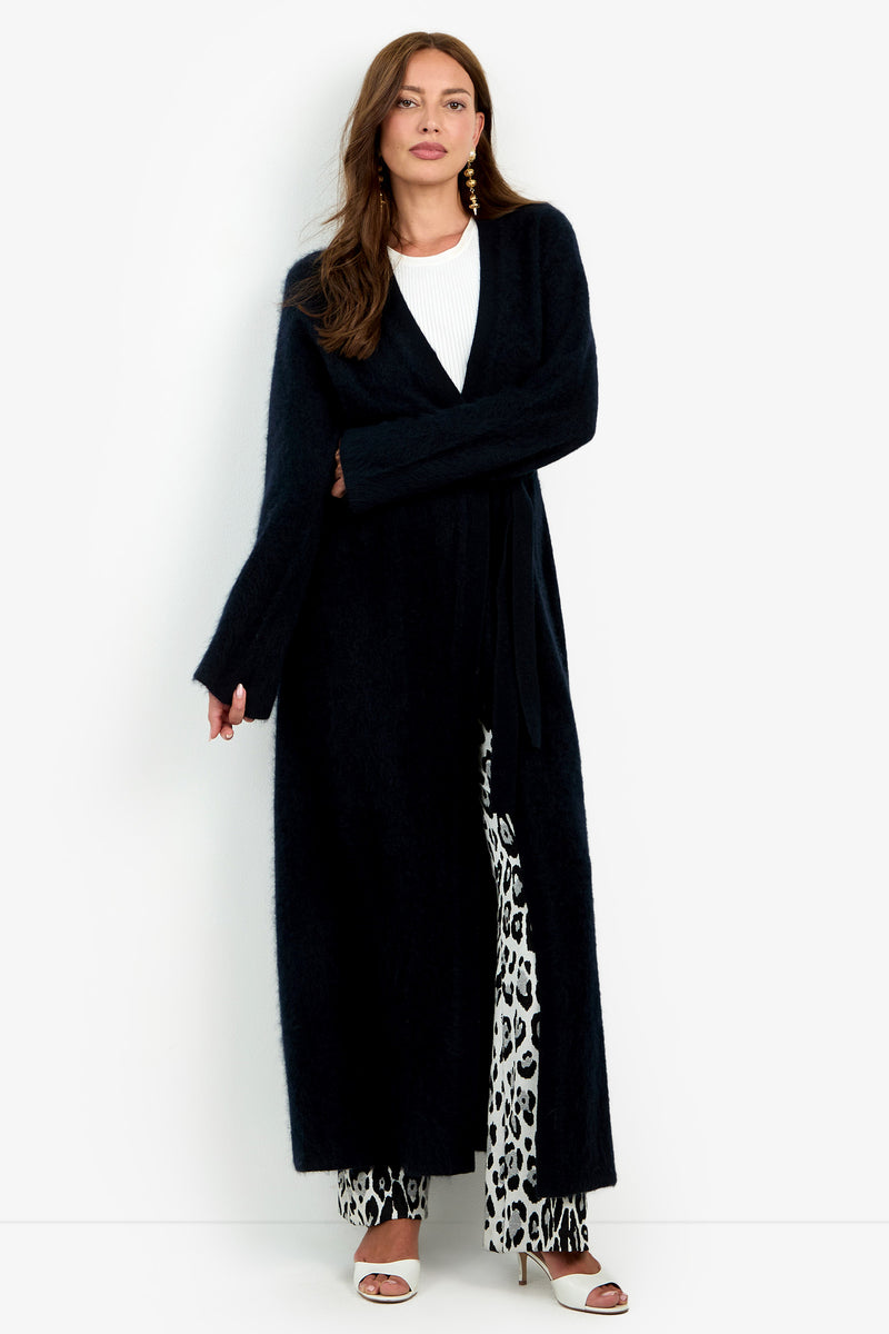 A woman wears a long, open black Carducci Cardigan over a white top and black-and-white patterned trousers, paired with white heeled sandals.