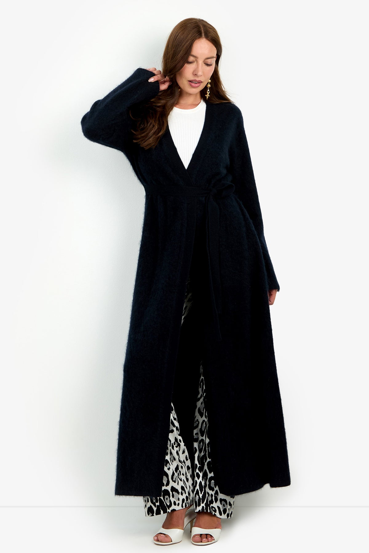 A woman wears a Carducci Cardigan, a long, open-front black piece with a tie waist, over a white ribbed top and black and white patterned trousers.