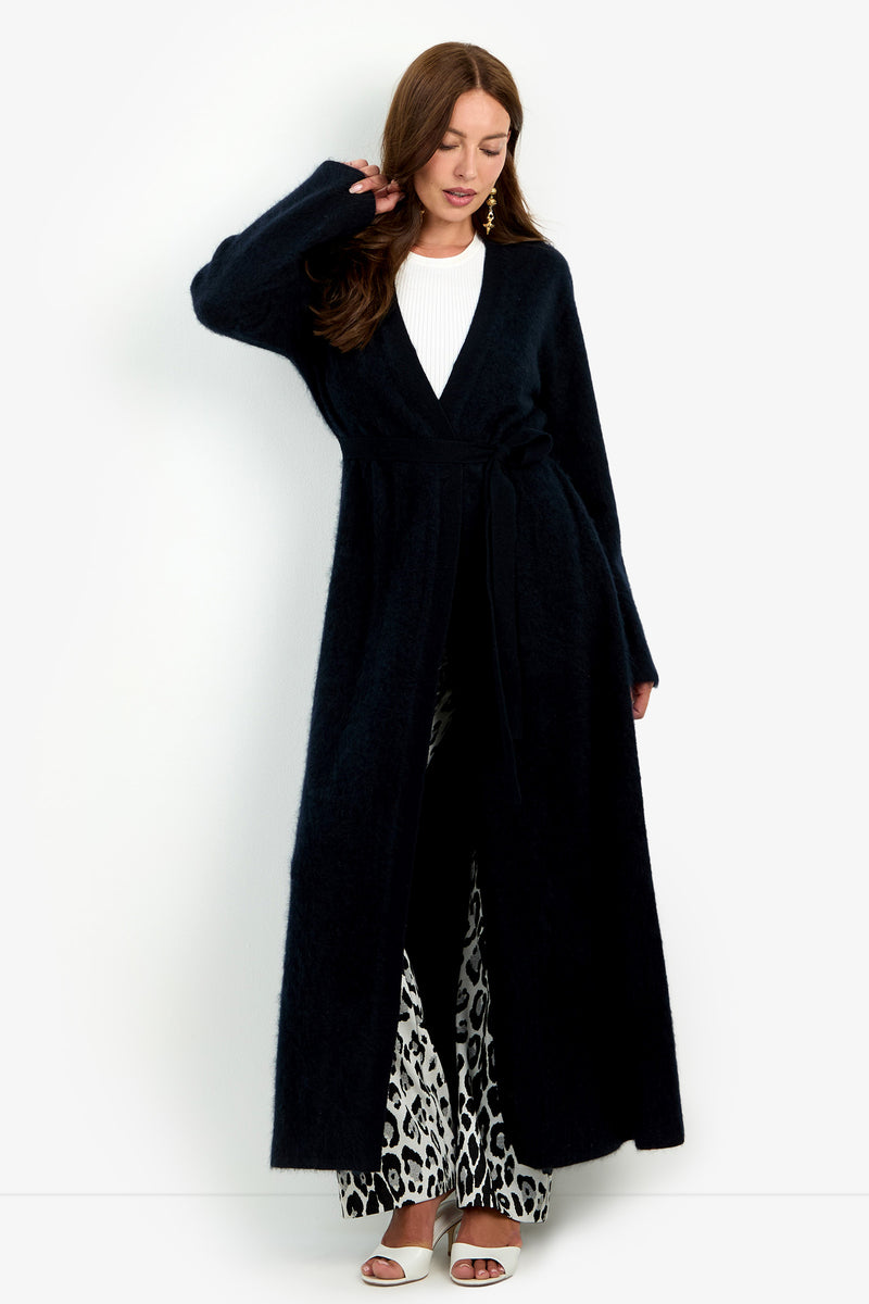 A woman wears a Carducci Cardigan, a long, open-front black piece with a tie waist, over a white ribbed top and black and white patterned trousers.