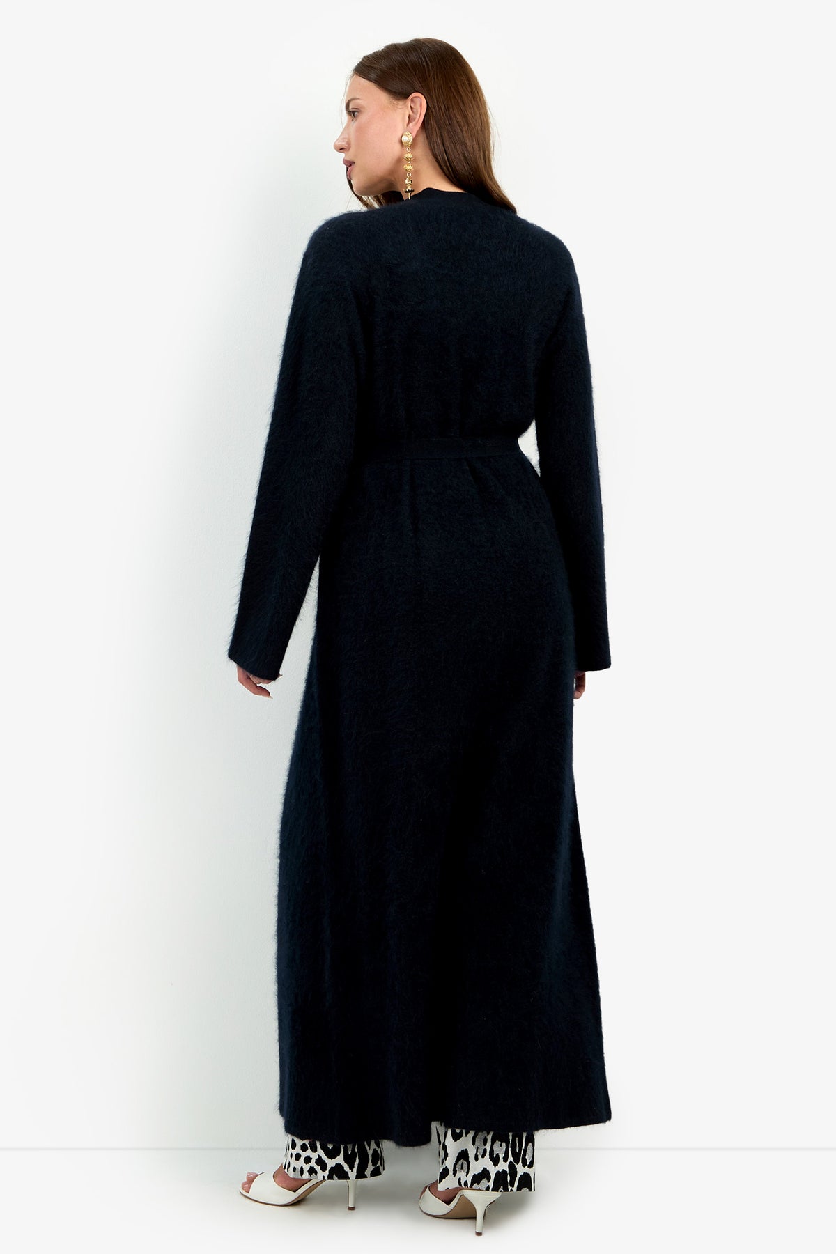 A person seen from behind wears a black, belted Carducci Cardigan over leopard-print pants and white heeled sandals.