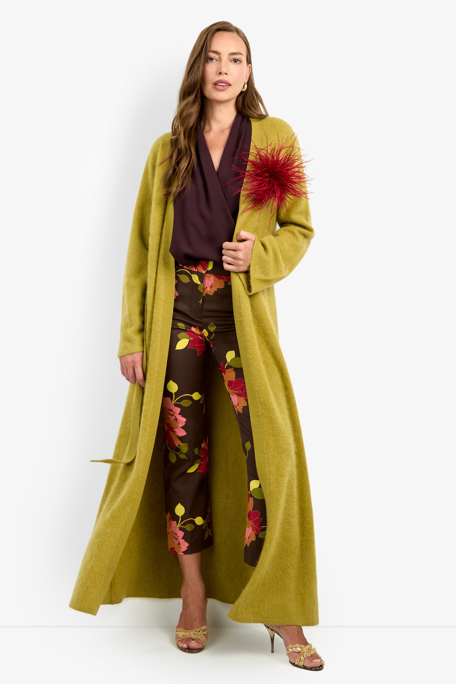 A woman models the CARDUCCI CARDIGAN, a long yellow-green coat with a large red flower detail, styled with a burgundy blouse, brown floral pants, and yellow heeled sandals. She stands against a white background.