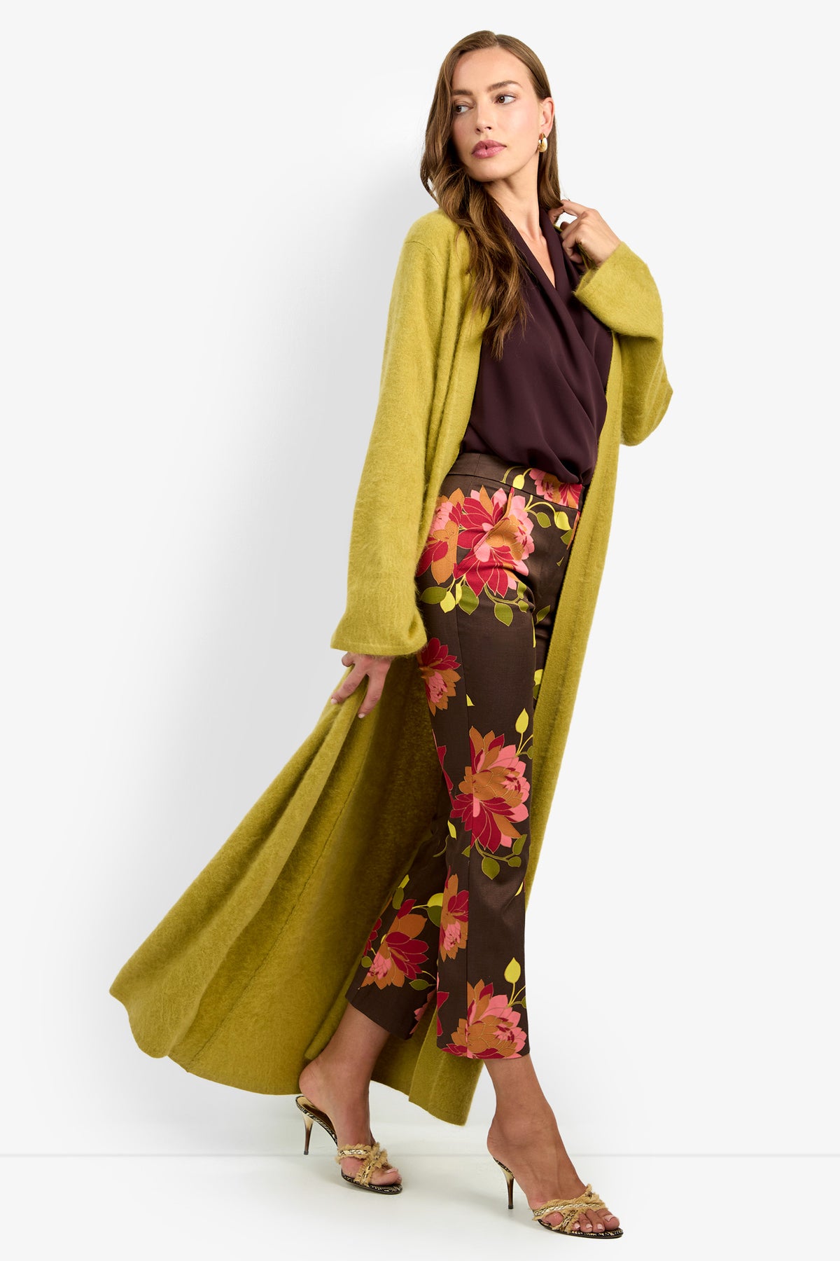 Wearing the CARDUCCI CARDIGAN, a woman poses in a long yellow-green piece layered over a burgundy blouse and floral print pants, paired with heeled sandals. She gazes to the side, relaxed and poised against a white background, one hand gently holding her cardigan open.