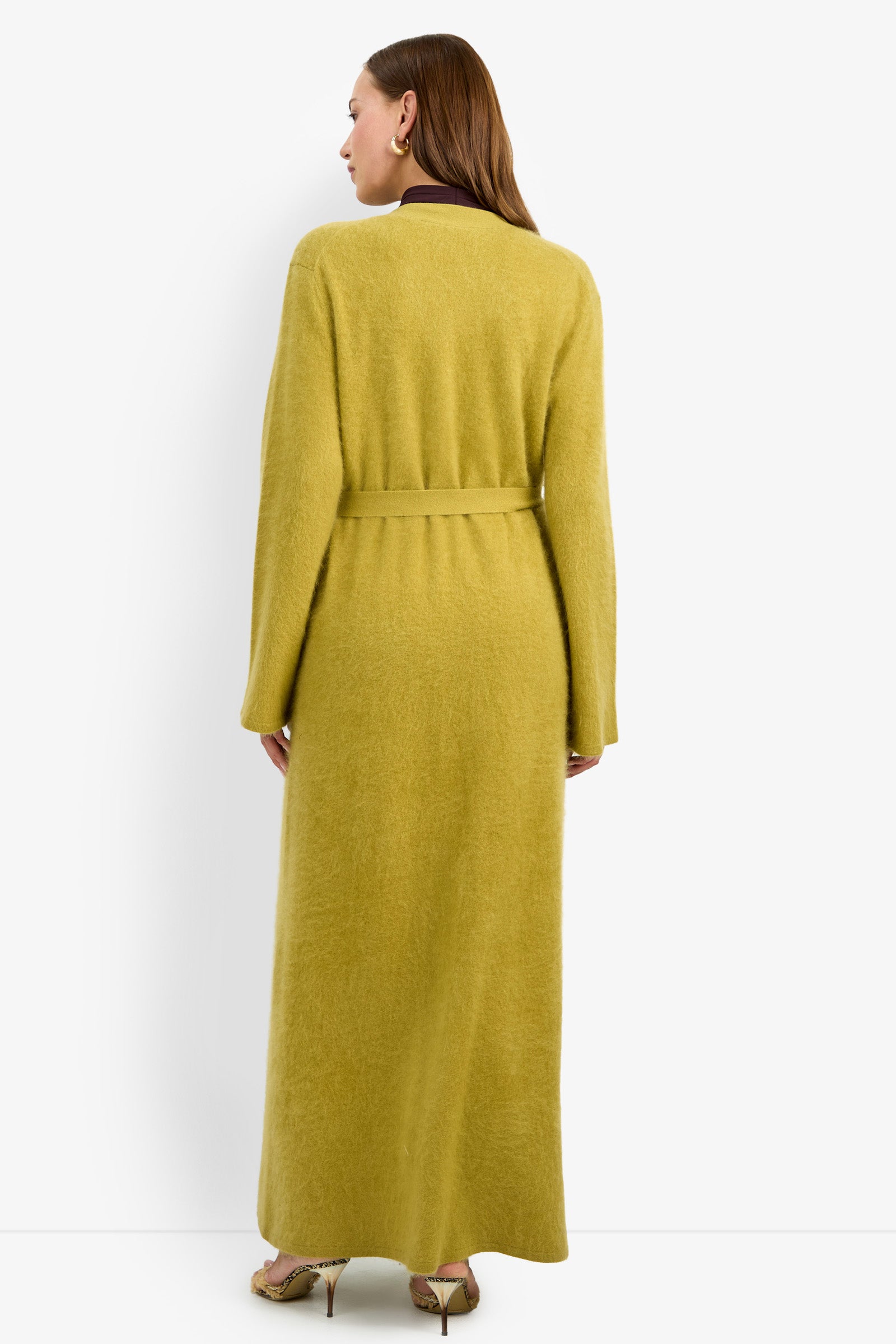 A woman faces away, wearing the CARDUCCI CARDIGAN—a long, yellow-green belted wool coat—paired with high-heeled shoes, and standing against a plain white background.