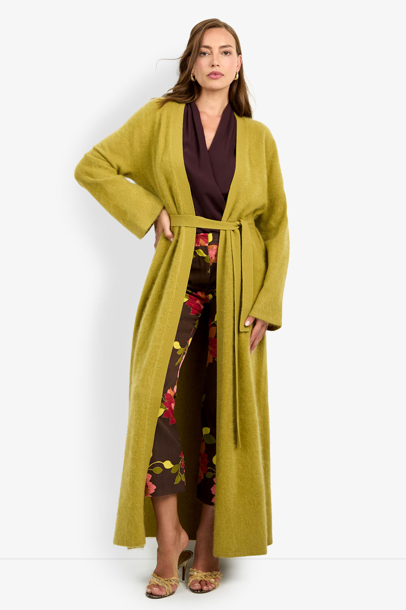 Woman wearing the CARDUCCI CARDIGAN, a long, light green belted coat, over a dark brown top and floral pants, standing against a plain white background.