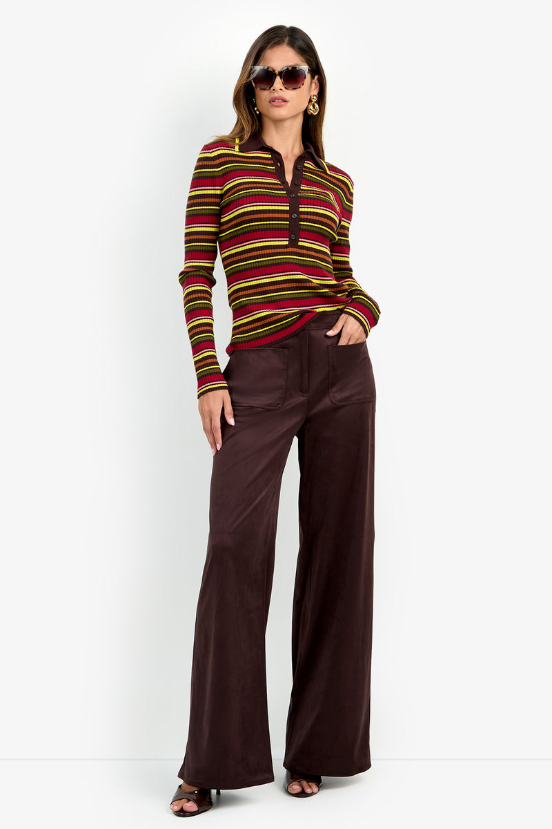 Woman wearing sunglasses, gold hoop earrings, the Hayworth Polo—a striped long-sleeve shirt in red, yellow, and brown—and wide-leg dark brown pants with open-toe heels, standing against a white background.