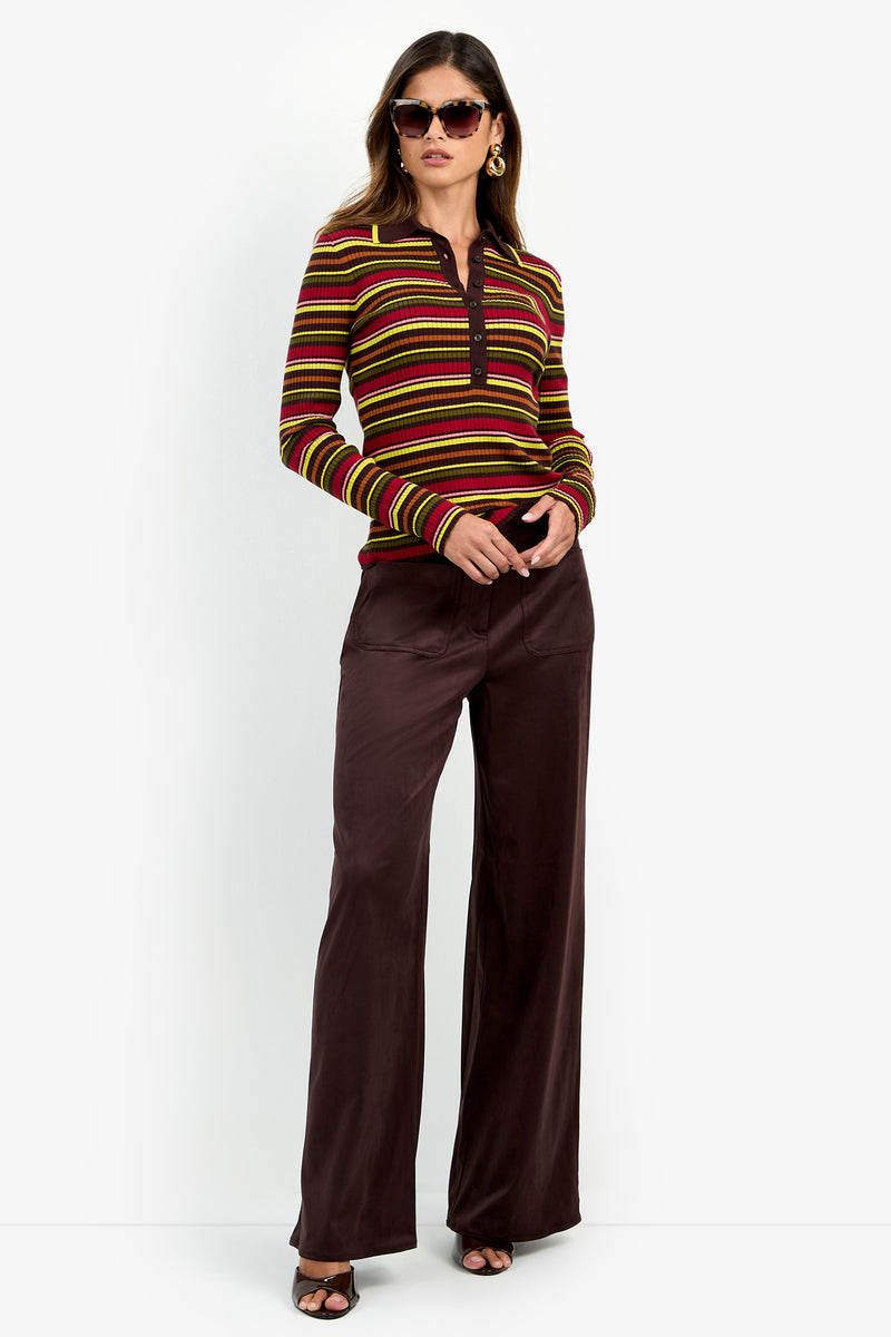 A woman wearing sunglasses and the Hayworth Polo—a striped long-sleeve top in red, green, and yellow—paired with high-waisted brown wide-leg pants, stands with her hands in her pockets against a plain white background.