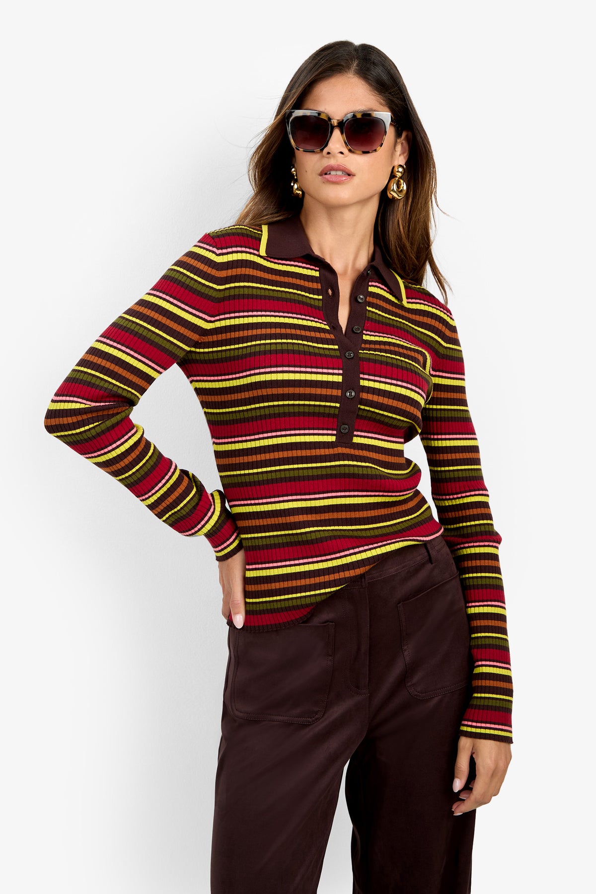 A woman wearing sunglasses, gold earrings, and the Hayworth Polo—a long-sleeved, striped collared shirt in shades of burgundy, yellow, and red—paired with dark pants poses with one hand on her hip against a plain white background.