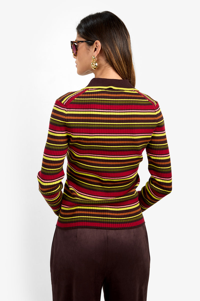 A woman with long brown hair, wearing sunglasses and gold earrings, faces away. She wears the Hayworth Polo—a ribbed, long-sleeve top featuring horizontal stripes in red, yellow, brown, and green—paired with dark brown pants.