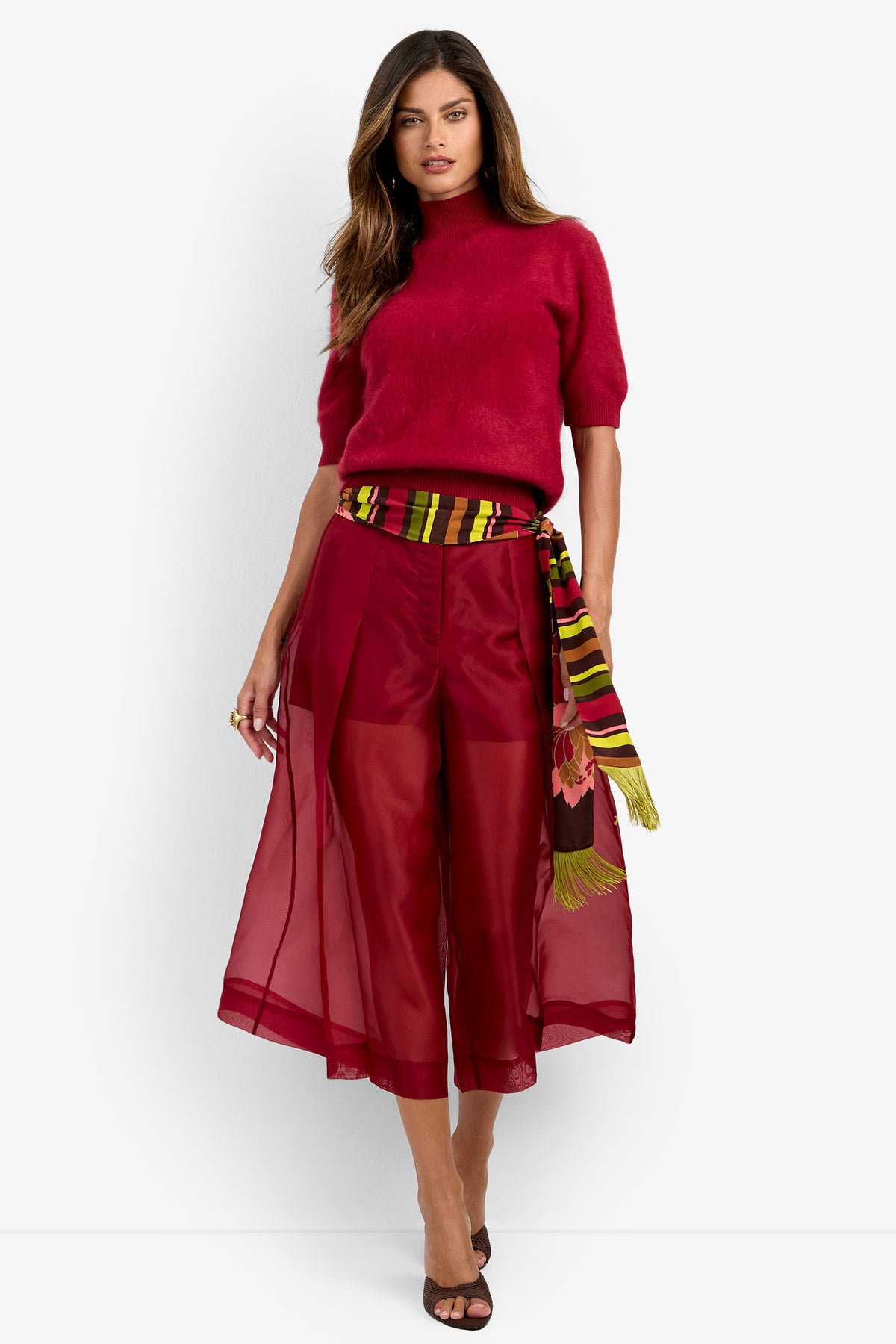 Woman wearing the Narni Short Sleeve Pullover in red, sheer wide-leg red pants, a colorful striped scarf belt, and black heels, standing against a white background.