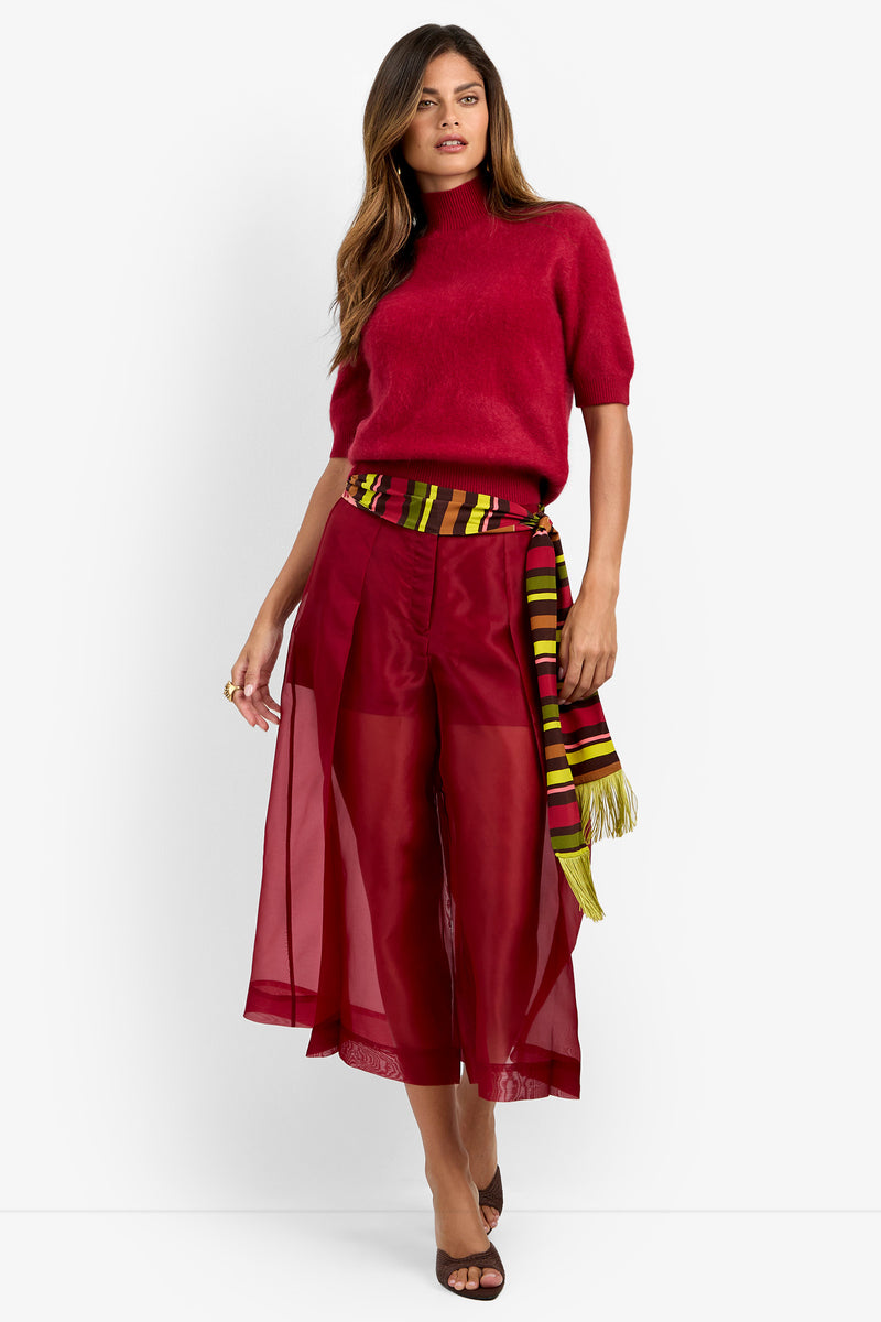 A woman stands against a plain white background wearing the Narni Short Sleeve Pullover in red, paired with red wide-leg sheer pants and brown heels. She has a striped yellow, green, and burgundy scarf tied at her waist.