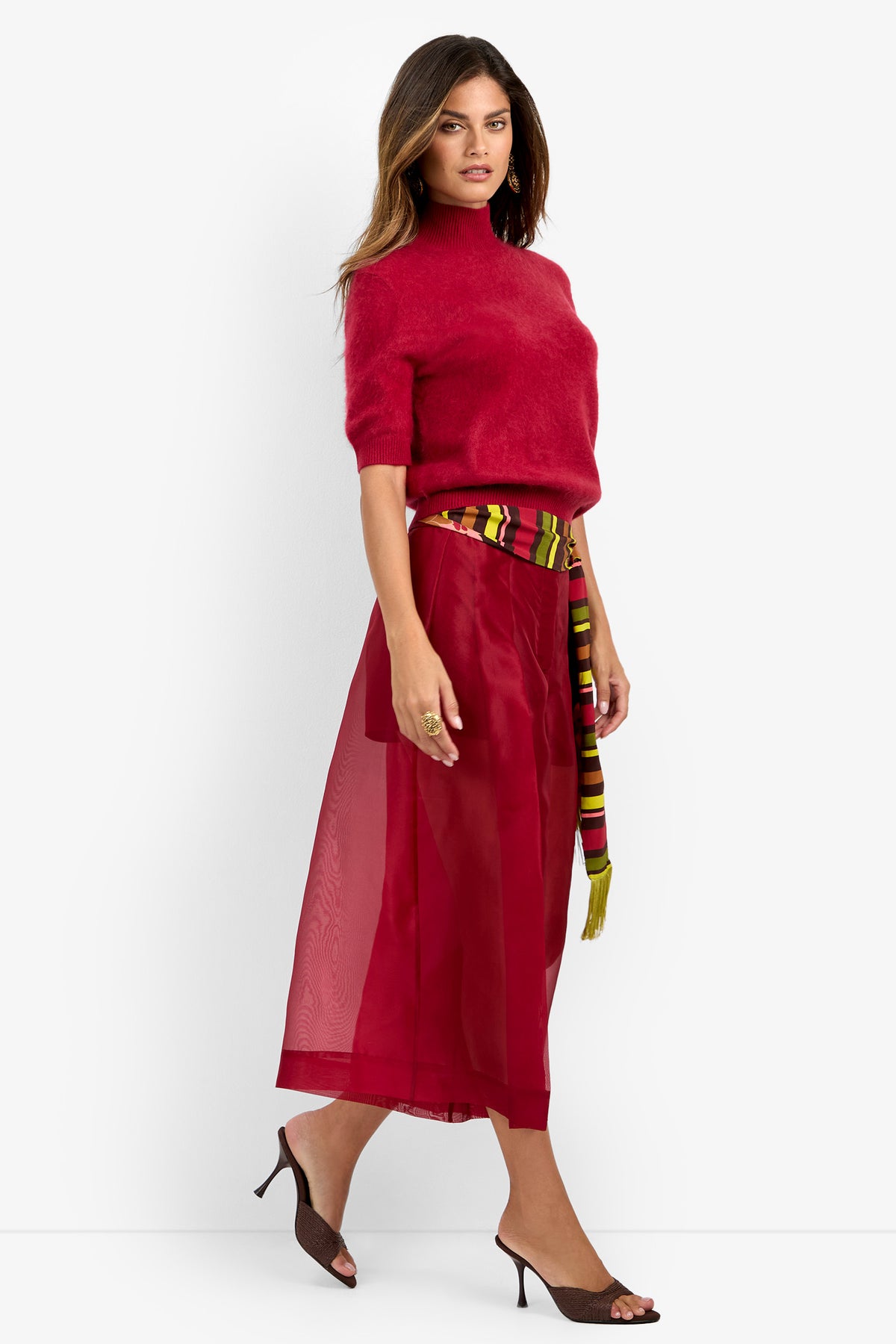 Woman wearing the Siena Pant with a red short-sleeve turtleneck sweater, sheer red midi skirt, multicolor sash belt, and brown heeled mules, walking against a plain white background.