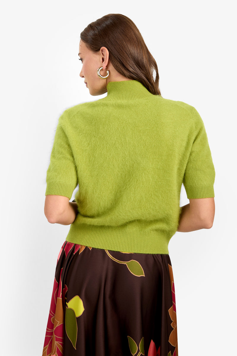 A woman with medium-length brown hair and gold hoop earrings faces away, wearing the Narni Short Sleeve Pullover in fuzzy lime green and a brown skirt featuring large, colorful floral patterns. The background is plain white.