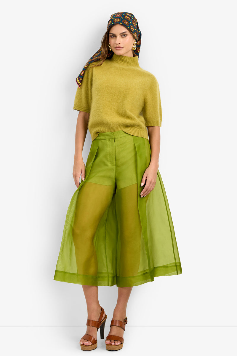 Wearing a mustard short-sleeve sweater, brown platform sandals, and a patterned headscarf, the woman stands against a plain background in the Siena Pant—sheer green wide-leg pants layered over shorts.