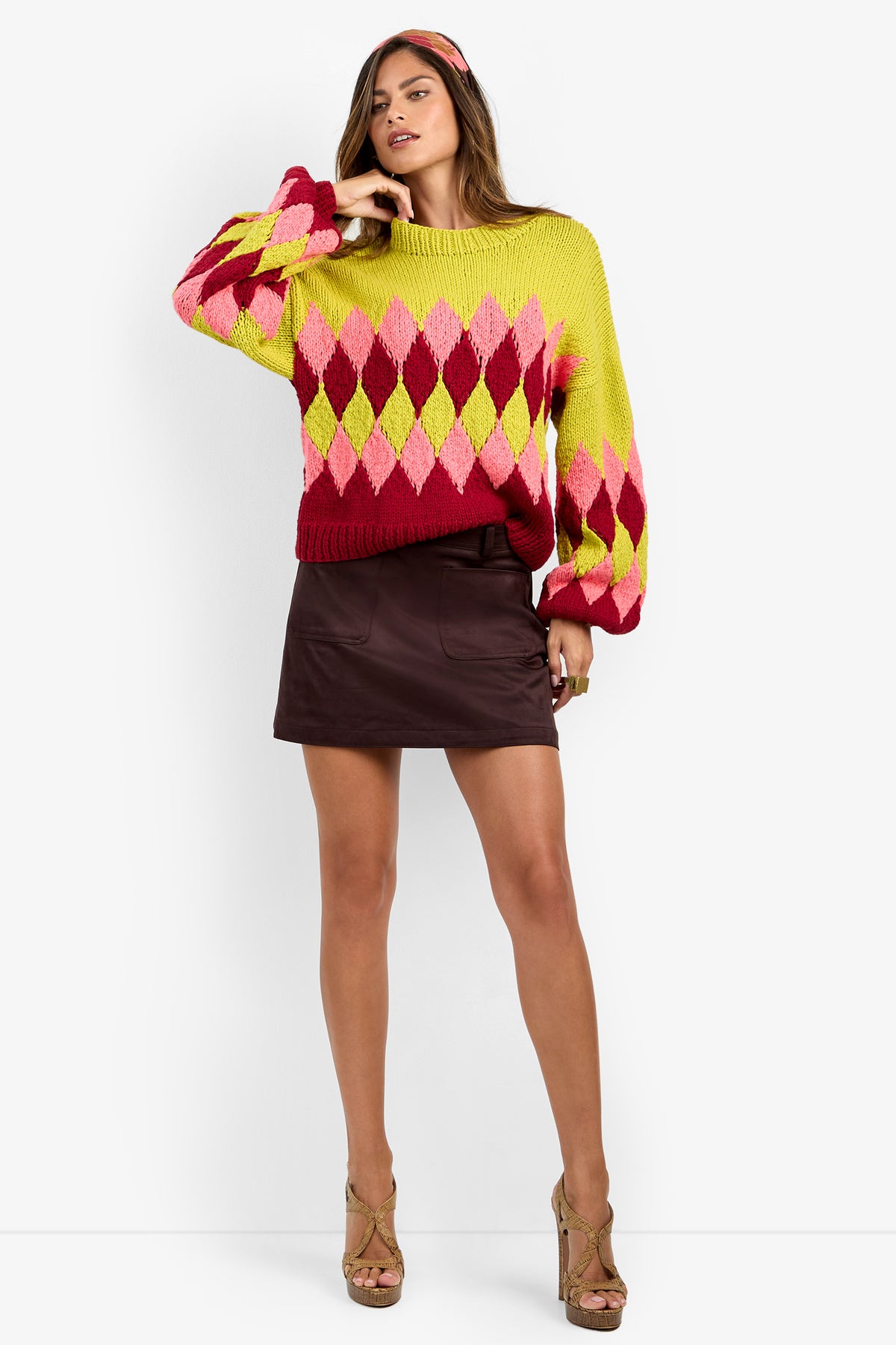 A woman stands against a white background, wearing the Cavallari Sweater—a chunky knit featuring a yellow, pink, and red diamond pattern—paired with a dark brown mini skirt, tan high-heeled sandals, and a matching headband.