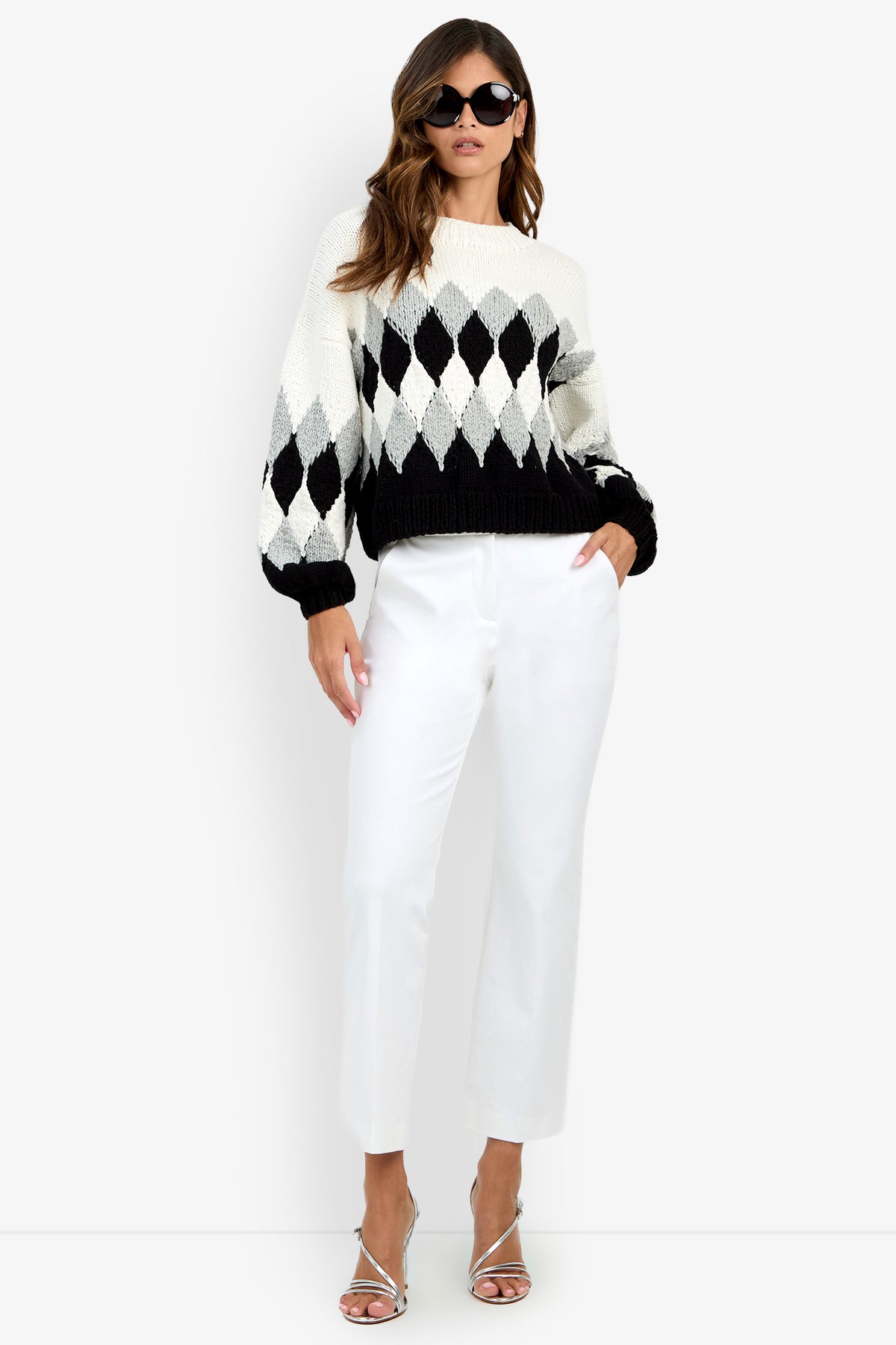 A person wearing a cream and black argyle Cavallari Sweater, white trousers, and metallic heeled sandals.