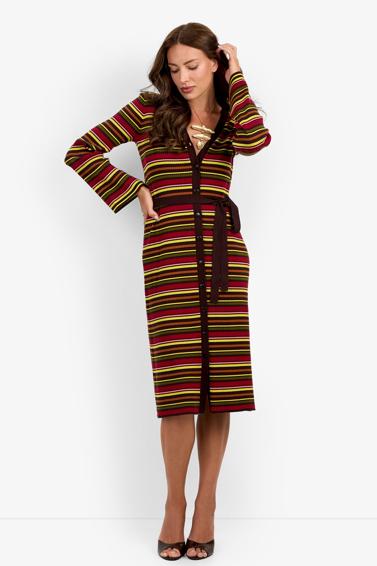 A woman with long brown hair stands with one hand touching her hair and looking down, wearing the Mercer Sweater Dress—a long-sleeve, belted dress featuring red, yellow, and black stripes—paired with black open-toed heels.
