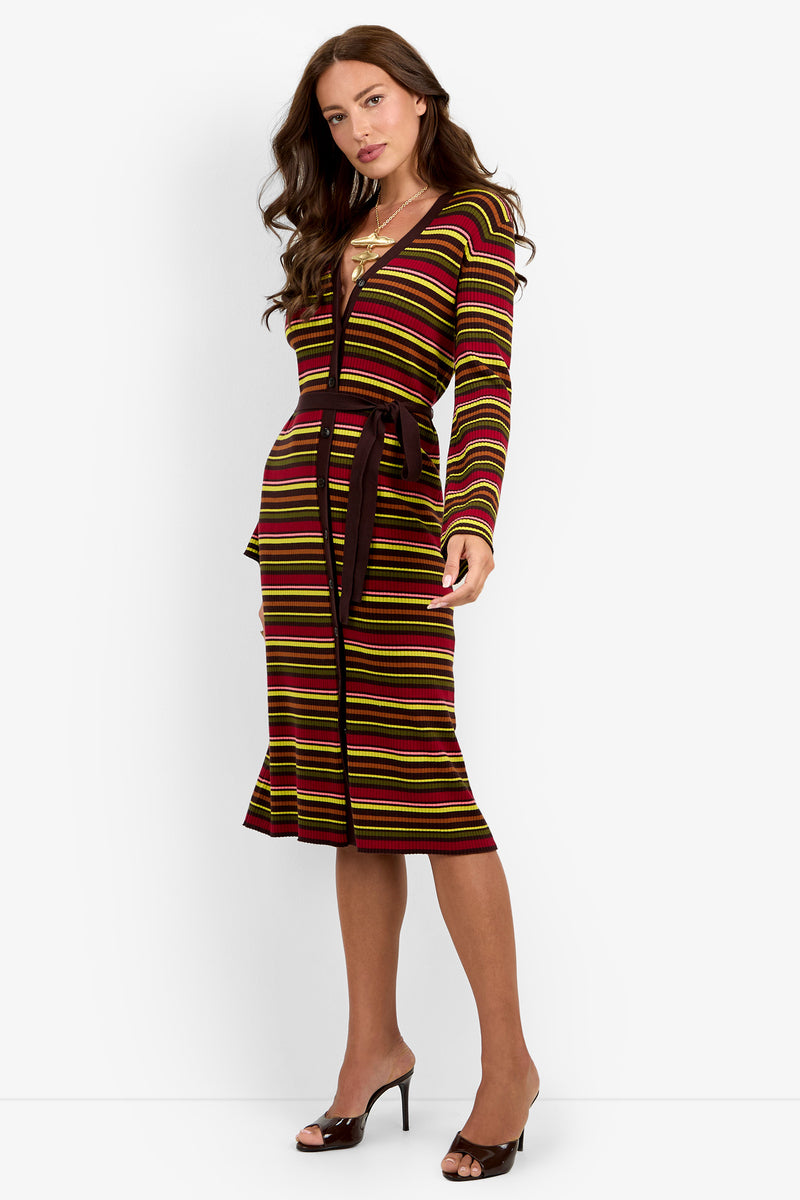 A woman is wearing the Mercer Sweater Dress, a fitted, knee-length dress with long sleeves and a V-neck, featuring horizontal stripes in brown, yellow, red, and green. The dress includes a matching belt and she completes the look with black open-toe heels.