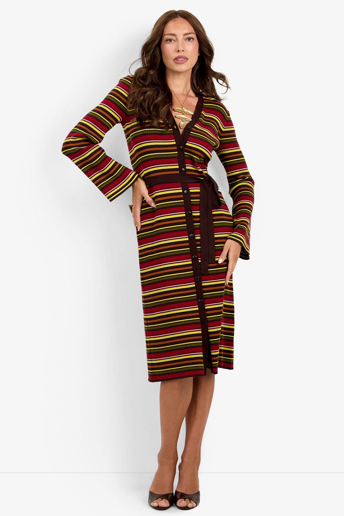 A woman poses against a plain white background, standing with one hand on her hip and the other touching her hair, wearing the knee-length Mercer Sweater Dress featuring long sleeves, multicolored stripes, and a dark belt.