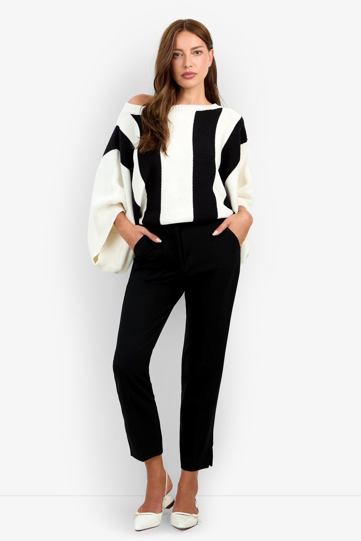 A woman models the black-and-white striped, off-the-shoulder Horizon Sweater with wide sleeves, paired with slim black trousers and white mules.