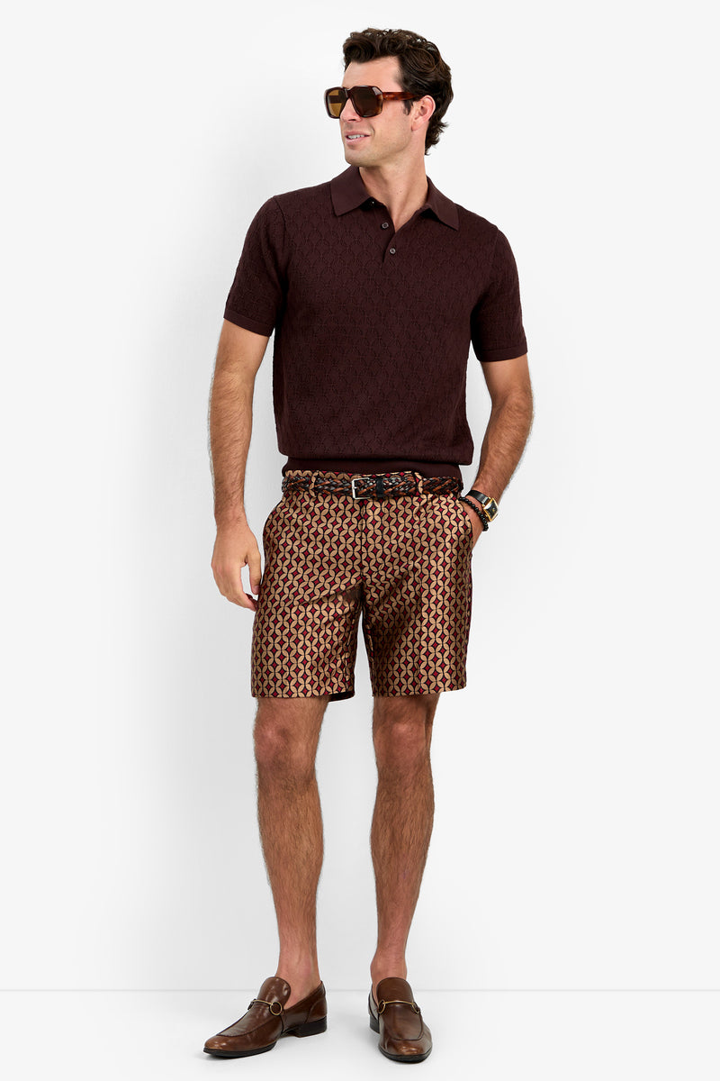 A man wearing sunglasses, the Waylen Polo in maroon, patterned shorts, a belt, and brown loafers stands against a white background.