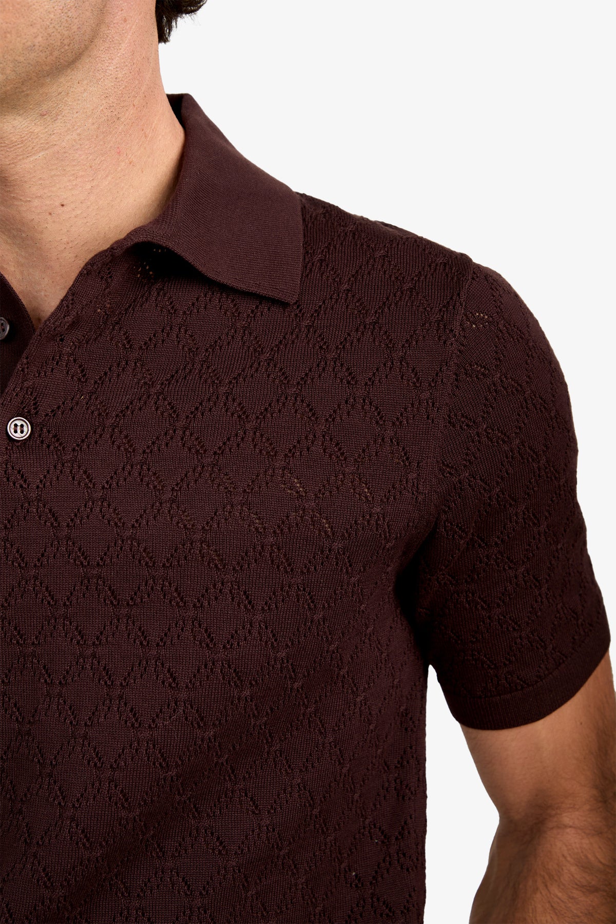 A close-up of a person wearing the Waylen Polo—a textured, dark brown short-sleeve shirt featuring a subtle diamond pattern. Only the shoulder, chest, and part of the face are visible.