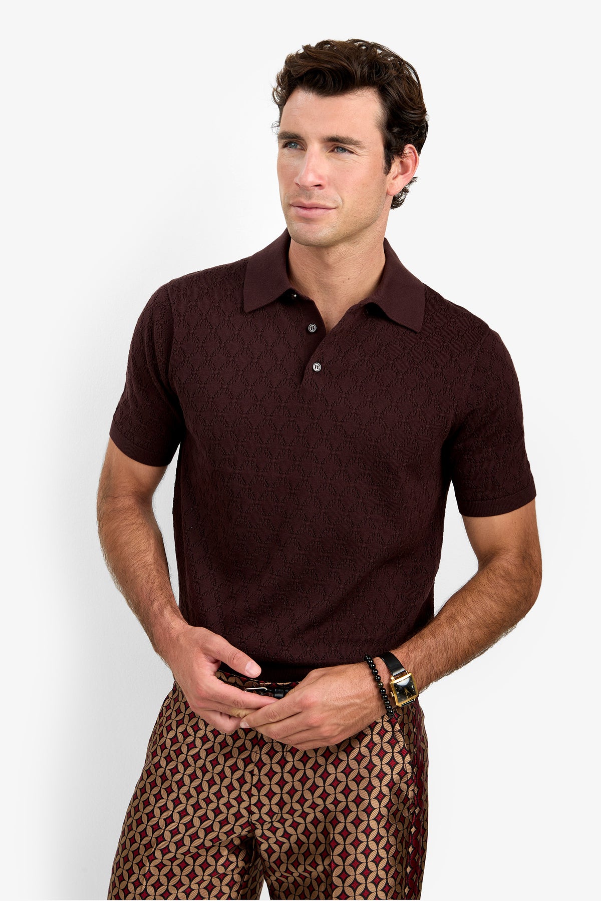 A man stands against a white background, wearing the short-sleeve maroon Waylen Polo and patterned pants. He has short brown hair, is looking slightly to the side, and rests his hands at his waist.