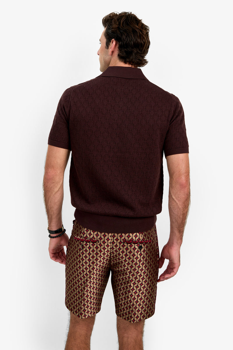 A man stands facing away, wearing the Waylen Polo—a short-sleeved dark burgundy shirt—paired with patterned geometric shorts, against a plain white background.