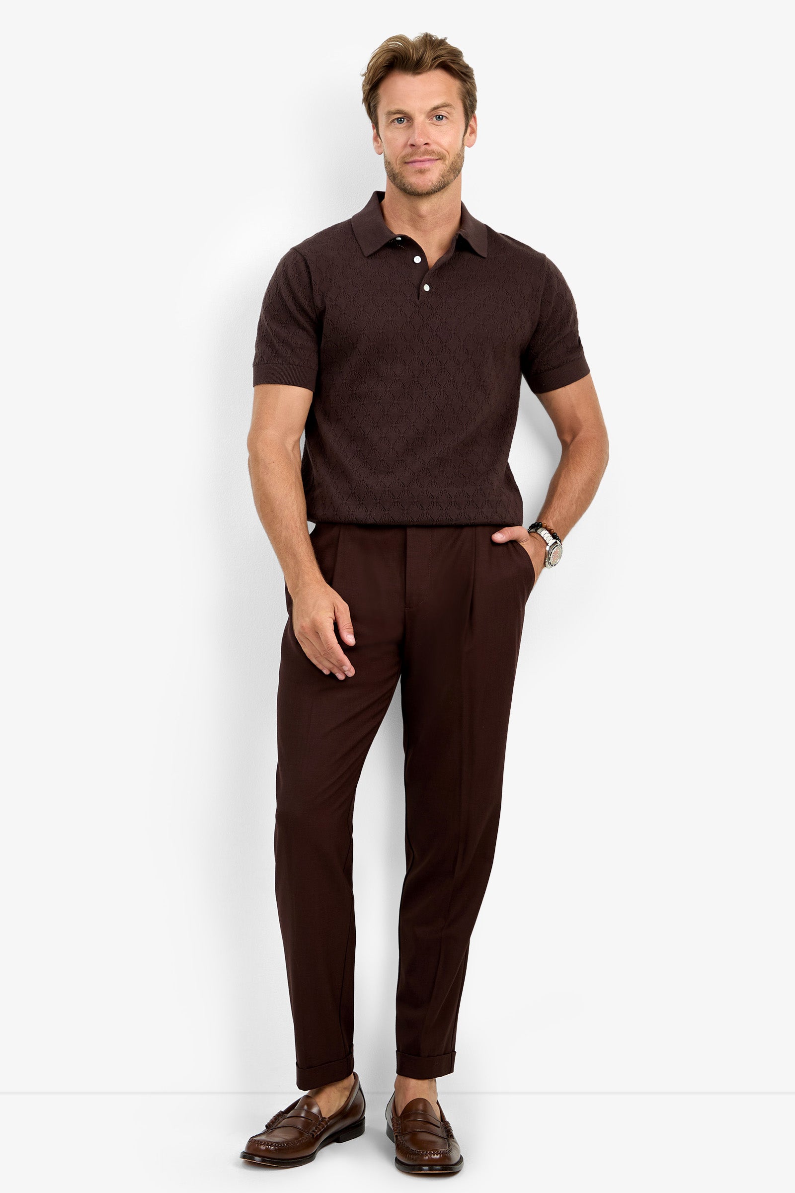 A man stands against a white background wearing the WAYLEN SS POLO, paired with brown pleated pants and brown loafers. He has one hand in his pocket and is smiling slightly.